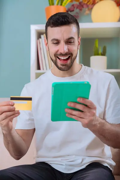 Neşeli bir adam kanepede oturup internetten alışveriş yapıyor. Her ikisi de dijital tablet ekranına heyecanla bakarken adamın elinde bir kredi kartı var. Dikey