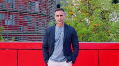 Ciddiyim. Spor ceketli olgun bir adam ciddi ciddi kameraya bakar. Şehir ortamında durur. Arkaplanda modern mimari ve yeşillik, profesyonel ve odaklanmış bir tavır sergileniyor