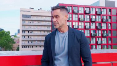 Ceketli olgun bir adam çatıda duruyor, gülümsüyor ve baş parmak işareti yapıyor. Canlı kent ortamında, olumlu ve güveni yansıtan kırmızı ve siyah binalar yer alıyor..