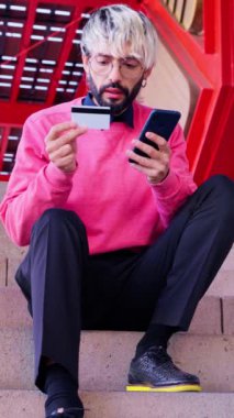 Saçları ağarmış ve gözlüklü modaya uygun genç bir adam elinde kredi kartı ve akıllı telefonuyla merdivenlerde oturuyor. Parlak pembe bir kazak giyen o, modern bir kentsel ortama odaklanmış görünüyor.. 