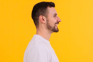 Beyaz tişörtlü neşeli bir gencin yan profili canlı sarı bir arkaplan ile yan profilde yer alıyor. Gülüşü ve sıradan görünüşü pozitif ve rahat bir hava yaratıyor.. 