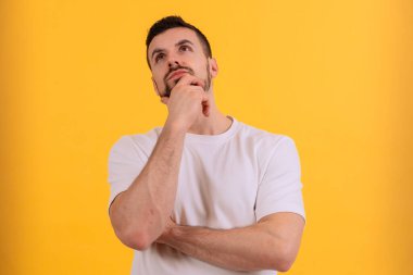  Beyaz gömlekli bir adam düşünceli bir şekilde elini şakağına koyuyor. Parlak sarı arka plan enerji ekler, bu görüntüyü düşünce, yaratıcılık veya karar verme mekanizması için ideal kılar.