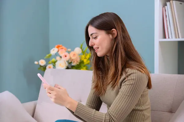 Genç bir kadın rahat bir koltukta oturuyor, akıllı telefonunu kullanırken gülümsüyor. Arka planda hafif ışıklandırma ve çiçek dekoru olan ev dekoru sakin ve rahat bir atmosfer yaratıyor.. 