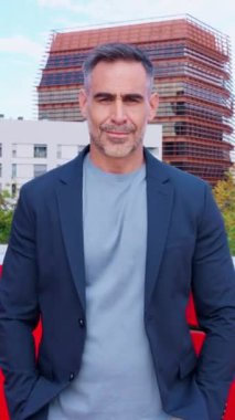 Ciddiyim. Spor ceketli olgun bir adam ciddi ciddi kameraya bakar. Şehir ortamında durur. Arkaplanda modern mimari ve yeşillik, profesyonel ve odaklanmış bir tavır sergileniyor