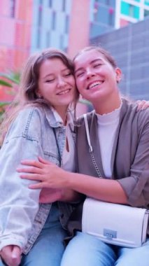  Açık havada yan yana duran ve kameraya içten bir şekilde gülümseyen iki neşeli genç kadının yakın plan fotoğrafı. Birinin uzun kahverengi saçları var, diğerinin de saçları arkadan bağlanmış. Güneş ışığı ağaçların arasından süzülür.