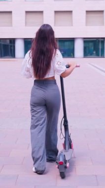 Uzun siyah saçlı genç bir kadın modern bir şehir meydanında elektrikli scooterın yanında yürüyor. Beyaz bir bluz ve gri bir pantolon giyiyordu. Kameradan uzaklaştı. Sıradan ve şık bir görünüm sergiliyordu. 