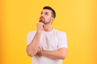  Beyaz gömlekli bir adam düşünceli bir şekilde elini şakağına koyuyor. Parlak sarı arka plan enerji ekler, bu görüntüyü düşünce, yaratıcılık veya karar verme mekanizması için ideal kılar.