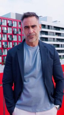 Neşeli, spor ceketli, gülümseyen olgun bir adam dışarıda oturur, baş parmağını kaldırır modern ortam yeşillik ve gösterişli mimari içerir, pozitif ve kendinden emin bir hava sergiler.. 