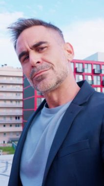 Ciddiyim. Spor ceketli olgun bir adam ciddi ciddi kameraya bakar. Şehir ortamında durur. Arkaplanda modern mimari ve yeşillik, profesyonel ve odaklanmış bir tavır sergileniyor