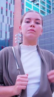 Kendine güvenen, şık genç bir kadın modern bir şehirde rahatça poz veriyor. Gri bir ceket giyiyor, beyaz bir bluz ve kot pantolon giyiyor, bir arka fonda duruyor. 
