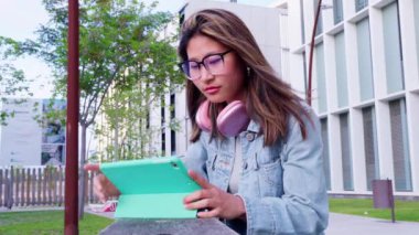 Gözlük ve kulaklık takan genç bir kadın dışarıda oturuyor ve tablete odaklanıyor. Binalar ve yeşillik ile modern ortam teknoloji, öğrenme ve kentsel yaşam tarzını ön plana çıkarıyor.