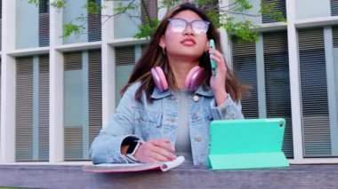 Gözlüklü ve kulaklıklı neşeli genç bir kadın dışarıda otururken telefonla konuşuyor. Tableti ve not defteri modern, teknoloji meraklısı ve üretken bir yaşam tarzını vurguluyor..