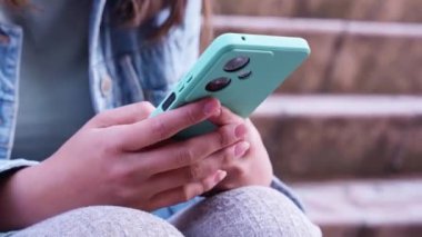  Açık havada otururken turkuaz akıllı telefon tutan birinin yakın çekimi. Kişi kot ceket giyer ve açık gri pantolon giyer. Sıradan, kentsel bir ortamda cihaz ekranına odaklanır.. 