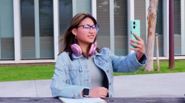 Kot ceketli ve gözlüklü gülümseyen genç kadın turkuaz akıllı telefonuyla selfie çekiyor. Boynunda pembe kulaklıklar var ve modern şehir parkında bir bankta oturuyor.. 