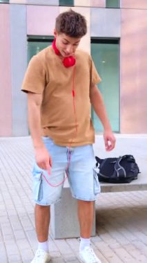 Genç bir adam. Günlük kıyafetler ve kırmızı kulaklıklar takan genç bir adam şehir merkezinin dışında duruyor, gülümsüyor ve yerdeki bir şeyi işaret ediyor. Sırt çantası yakınlardaki bir bankta duruyor, rahatlığı arttırıyor.