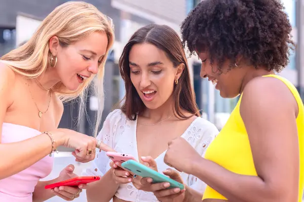 Üç neşeli genç kadın kırmızı bir akıllı telefonun etrafında toplanıyor, heyecanla tepki veriyorlar. Gelişigüzel giyiniyorlar, modern bir şehir ortamında dışarıda duruyorlar, birlikte eğlenceli bir anın tadını çıkarıyorlar..