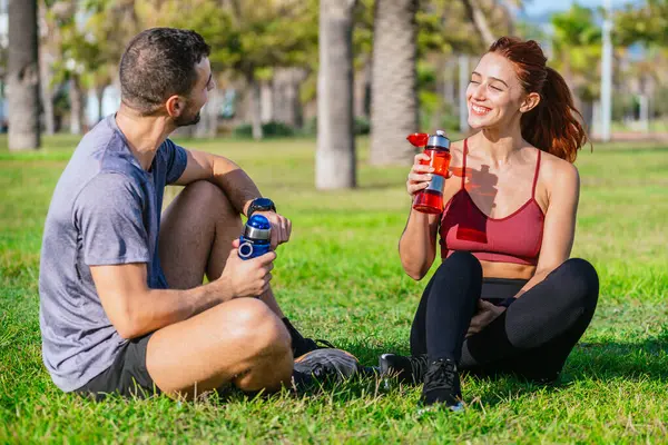 Formda bir erkek ve bir kadın spor yaparken çimlerin üzerinde oturur, güneşli bir parkta gülümser ve idmandan sonra su şişeleriyle su içerler..
