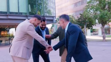 Takım elbiseli dört profesyonel iş adamından oluşan bir grup modern bir ofis binasının önünde takım çalışması ve birlik jesti olarak gülümseyip el ele tutuşuyorlar.