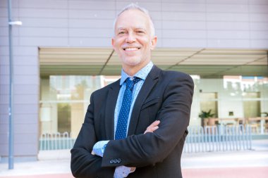 Takım elbiseli profesyonel bir işadamı kollarını kavuşturup modern bir ofis binasının önünde kendinden emin bir şekilde gülümser..