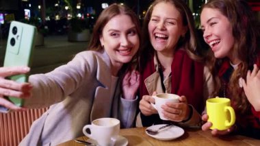 Üç gülümseyen kadın gece bir kafede selfie çekiyor, ellerinde kahve fincanları ve mutlu poz veriyorlar. Dışarıda çekilen eğlenceli ve neşeli bir an..