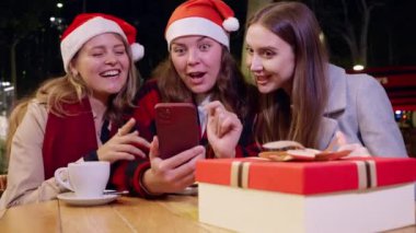 Noel Baba şapkası takan üç genç kadın gece açık havada akıllı telefona bakarken gülümsüyor. Şenlikli bir hediye kutusu ve kahve fincanı ahşap masada oturuyor.