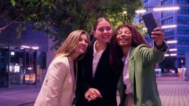 İş kıyafetleri içinde üç gülümseyen kadın gece şehir dışında bir yerde selfie çekerek şehir ışıkları altında bir anlık eğlence ve bağlantının tadını çıkarıyorlar.