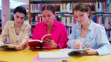 Üç genç kadın kütüphanede bir masada oturup kitap okumaya odaklanmış. Günlük kıyafetler giyiyorlar ve kütüphanede kitap okuyan renkli kitaplarla dolu raflarla çevrililer.. 