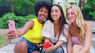 Üç genç kadın açık hava basamaklarında birlikte oturup pembe akıllı telefonlarıyla selfie çekerken gülümsüyorlar. Günlük yaz kıyafetleri giyiyorlar ve etrafları yeşilliklerle çevrili. Açık havada selfie çekiyorum..