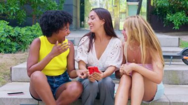 Üç neşeli genç bayan açık hava basamaklarında oturup gülüşüyor ve sohbet ediyorlar. Birinin elinde kırmızı bir akıllı telefon var. Yeşilliklerle çevrili günlük yaz kıyafetleri giyiyorlar.. 