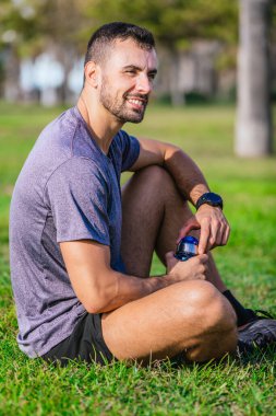Parktaki çimlerin üzerinde oturmuş elinde su şişesiyle gülümseyen atletik bir adam. Spor giyer, spor giyer ve dışarıda egzersiz yaptıktan sonra ara verir..