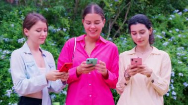 Üç genç kadın dışarıda duruyor, akıllı telefondaki bir şeye heyecanla tepki veriyorlar. Günlük giysiler giyiyorlar ve yemyeşil ile çevrililer. Smartphone Content Outdoor 'a tepki. 