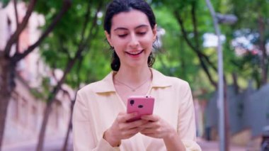  Genç bir kadın açık havada ahşap bir bankta pembe akıllı telefonunu kullanarak oturuyor. Açık renk bir bluz ve kot pantolon giyiyor. Odaklanmış ve rahat görünüyor..