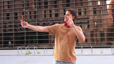 Gündelik kıyafetlerle gülümseyen genç adam akıllı telefonuyla selfie çekiyor, kulaklık takıyor ve modern cam bir binanın önünde poz veriyor.. 