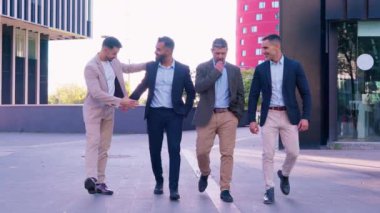İş dünyasından dört iş adamı resmi ve günlük giysiler içinde şehir ortamında birlikte yürüyorlar, rahat bir anda sohbet ve arkadaşlıkla iç içeler.