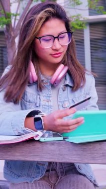 Kulaklıklı genç bir kız öğrenci, açık havada elinde dijital tablet ve kredi kartıyla online alışveriş yapıyor. Konsept: Çevrimiçi alışveriş.