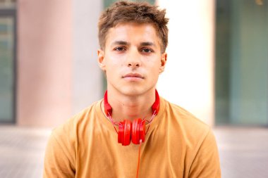 Genç adam boynuna kırmızı kulaklık takan ve bronz gömlek giyen bir gencin portresi. Açık hava ortamında kısa kahverengi saçlı nötr bir ifade.