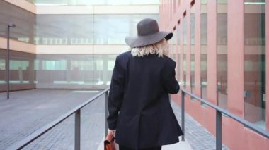 Geniş kenarlı şapkası ve paltosu olan şık bir kadın cep telefonuyla konuşurken çağdaş bir şehir manzarasında yürüyor. Deri bir çanta taşıyor, zarafet ve profesyonellik yayıyor..