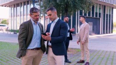 Resmi giyinmiş iki işadamı ciddi ifadeleri olan bir akıllı telefona bakarken, açık şehir ortamında işle ilgili konuları tartışırken, diğerleri arka planda sohbet ederken,