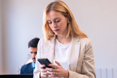 BusinesswFocused businesswoman in a beige blazer checking her smartphone in a modern office setting, with a male colleague working in the background clipart