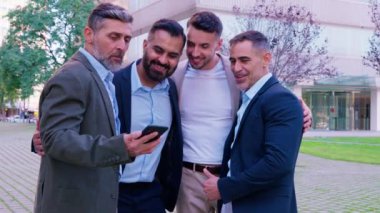 Resmi ve günlük giysiler içinde dört işadamı, güler yüzlü ve açık bir şehir ortamında grup selfie 'sine poz vererek neşeli ve rahat bir anı yakalar..