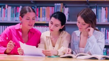  Kütüphanede bir masada oturan üç genç kadın, arka planda kitaplıklarla çevrili bir projede işbirliği yaparken belge ve kitapları tartışıyorlar. Bir kütüphanede. Yüksek kalite 4k görüntü