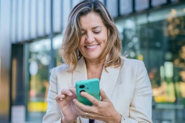 Gülümseyen orta yaşlı, bej ceketli, arka planda bulanık modern bir binayla dışarıda akıllı telefon kullanan mutlu bir kadın. 