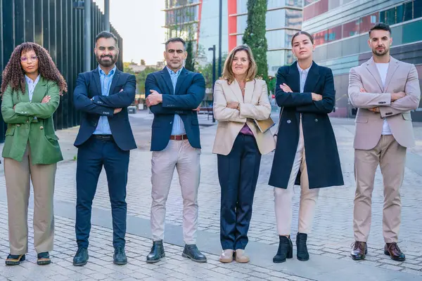  İş kıyafetleri içinde çeşitli profesyonellerden oluşan bir grup, arka planda modern binaların bulunduğu kentsel bir ortamda açık havada, kolları çaprazlanmış bir şekilde kuyrukta duruyorlar.. 