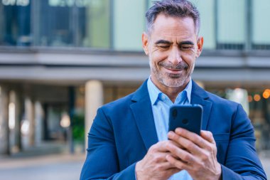 Smiling middle-aged businessman in a blue shirt holding and using a smartphone outdoors in a modern urban setting. clipart