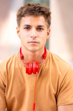 Genç adam boynuna kırmızı kulaklık takan ve bronz gömlek giyen bir gencin portresi. Açık hava ortamında kısa kahverengi saçlı nötr bir ifade.