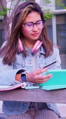 Kulaklıklı genç bir kız öğrenci, açık havada elinde dijital tablet ve kredi kartıyla online alışveriş yapıyor. Konsept: Çevrimiçi alışveriş.