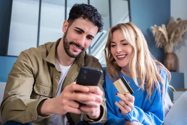 Keyifli bir çift rahat bir oturma odasında oturmuş, akıllı telefon ve kredi kartını online alışveriş için kullanıyorlar. Evdeki modern yaşam tarzı konsepti ve teknoloji.