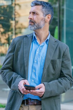 Sakallı, ceketli, mavi gömlekli, akıllı bir telefon tutan ve modern cam mimarisi olan şehir dışı bir çevreye bakan olgun bir adam..