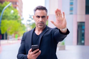 A mature businessman in a black suit holding a smartphone and making a stop gesture in an urban setting. Professional, determined expression, symbolizing authority and focus. clipart