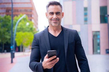 Mavi tişörtlü, gülümseyen orta yaşlı bir işadamı modern bir şehir ortamında akıllı telefon kullanıyor..